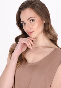 Világosbarna, ujjatlan felső V-nyakkivágással; sima, puha textúrájú; a modell keze az állán pihen, természetes sminkkel és hullámos hajjal.
