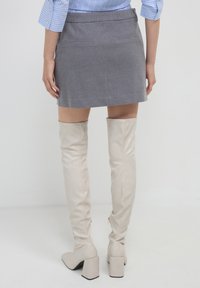 Frau in einem grauen Minirock, blau gestreiftem Langarmshirt und beigen kniehohen Stiefeln mit Absatz, stehend vor einem weißen Hintergrund.