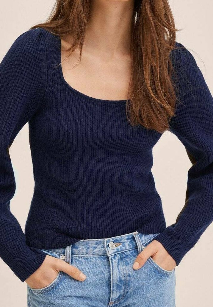 Femme portant un pull côtelé bleu marine ajusté à manches longues et un jean taille haute bleu clair, les mains dans les poches.