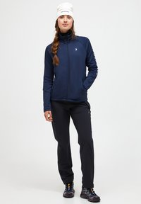 Navy Zip-Jacke aus glattem Stoff, kombiniert mit schwarzen Hosen; dazu eine weiße Mütze und Outdoorschuhe mit ausgeprägtem Profil.