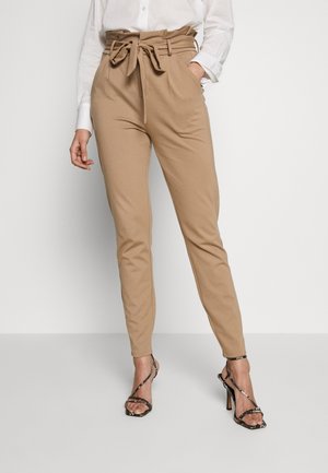 Vrouw met beige hoge taille broek vastgebonden met een riem, een wit overhemd en zwarte sandalen met bandjes en hoge hakken, staand tegen een effen achtergrond.