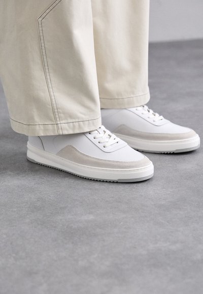 Hvide sneakers med en glat læderoverdel, akcentueret med beige ruskind, flade snørebånd og en tekstureret gummisål, sat sammen med lyse, vide bukser.