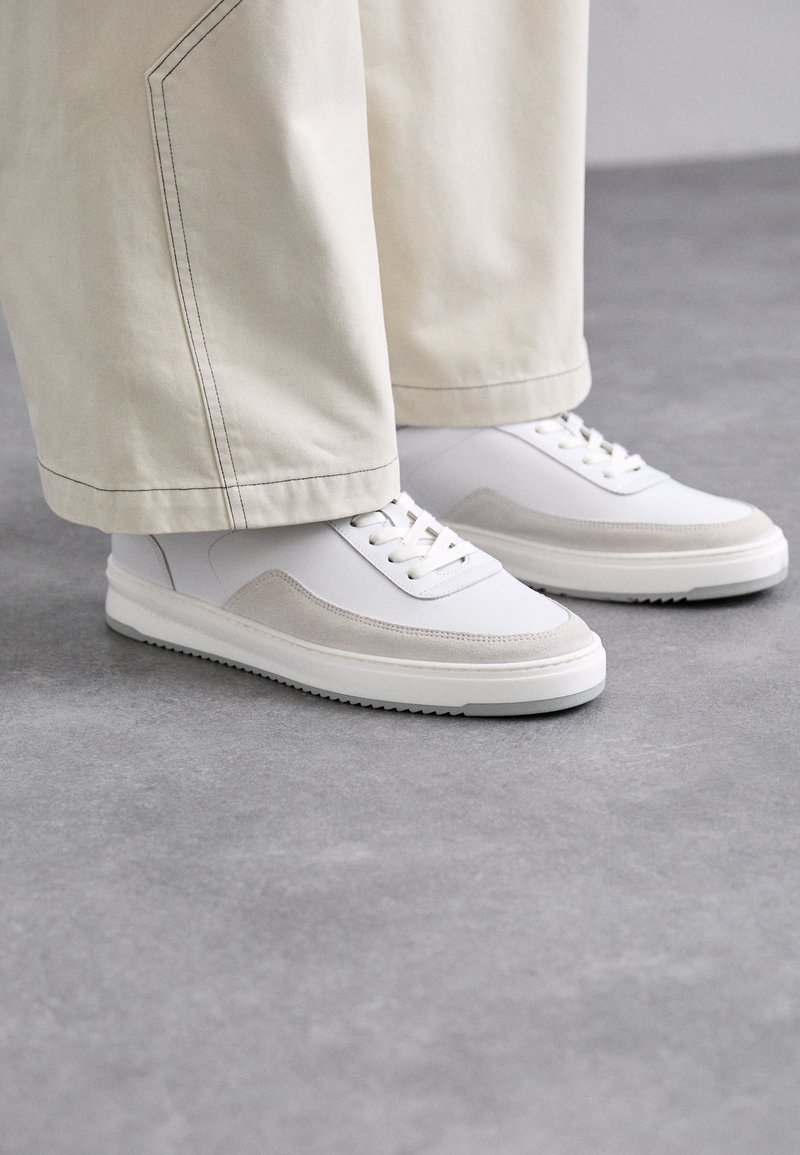 Baskets blanches avec une tige en cuir lisse, des accents en daim beige, des lacets plats et une semelle en caoutchouc texturée, associées à un pantalon large de couleur claire.
