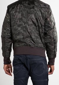Man i mörk kamouflage-bomberjacka och mörka jeans, sedd bakifrån mot en neutral bakgrund.