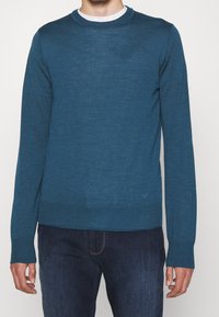 Blue crew-neck sweater made from a fine-knit material, featuring long sleeves and a ribbed hem, paired with dark denim jeans.