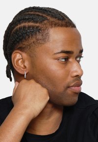 Profilansicht einer Person mit geflochtenem Haar, die einen silbernen Creolenohrring trägt. Der Hintergrund ist schlicht weiß, und sie stützt ihr Kinn auf ihre Hand.