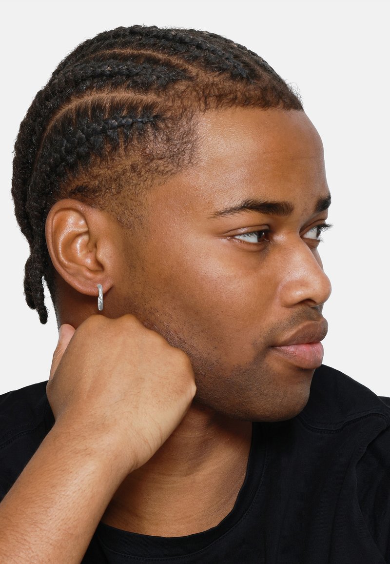 Profilansicht einer Person mit geflochtenem Haar, die einen silbernen Creolenohrring trägt. Der Hintergrund ist schlicht weiß, und sie stützt ihr Kinn auf ihre Hand.