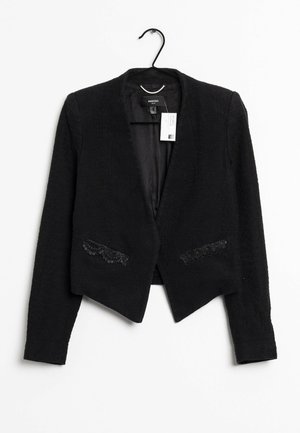 Blazer noir court à manches longues, poches avant bordées de dentelle, suspendu à un cintre noir sur fond blanc.