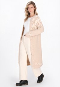 Lange beige cardigan met een textuur, gebreid ontwerp met een zigzagpatroon op de mouwen en een rij knopen aan de voorkant. Witte shirt en crèmekleurige broek.