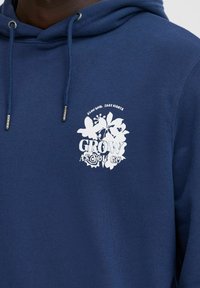 Sötétkék kapucnis pulóver, puha anyagból készült, fehér színű virágos grafikával és a "GROW AS YOU GO" felirattal a bal mellrészen.