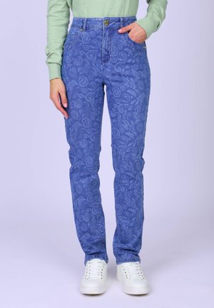 Femme portant un jean bleu à motifs cachemire, un haut à manches longues vert clair et des baskets blanches, avec une main dans la poche et l'autre détendue.