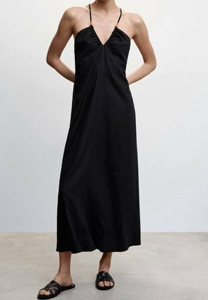 Persona con un vestido largo negro sin mangas con tirantes finos y cuentas de madera, combinado con sandalias negras planas de tejido entrelazado, de pie contra un fondo liso.