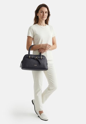 Femme en tenue blanche tenant un sac à main en cuir noir avec des accents dorés, debout avec une jambe croisée sur l'autre devant un fond neutre.