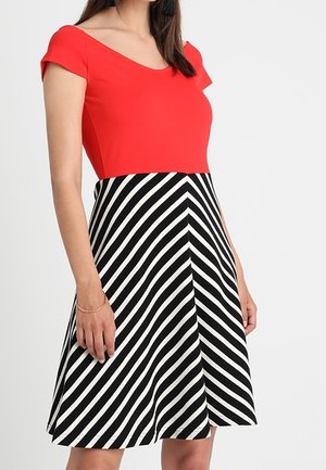 Frau trägt ein rotes tailliertes Oberteil mit kurzen Ärmeln und einen schwarz-weiß diagonal gestreiften Rock.