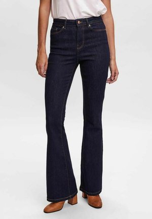 Zvonové tmavě modré džíny z denimu s viditelným šitím, vysokým pasem a mírným elastickým materiálem pro lepší přizpůsobení. V kombinaci s hnědými podpatky.