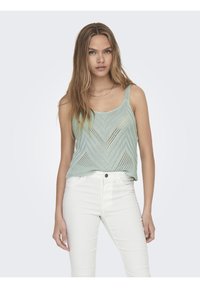 Débardeur en maille vert clair avec un motif en chevron, de larges bretelles et un design en tissage ouvert, associé à un pantalon blanc.