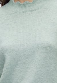 Pull tricoté vert clair doux avec un col montant côtelé festonné porté par une personne.