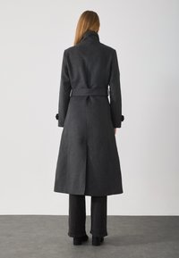 Long, dark gray coat with a high collar, belt at waist, and a split back. Made of textured fabric, fitted design, and finished with rolled cuffs.