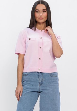 Jeune femme aux cheveux bruns lisses portant un haut rose clair à manches courtes boutonné et un jean taille haute bleu, posant devant un fond blanc.