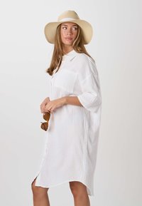 Femme portant une robe boutonnée blanche ample et un chapeau de paille à large bord, tenant des lunettes de soleil écaille de tortue, regardant vers sa gauche.