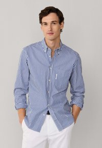 Camicia a righe con bottoni, caratterizzata da strisce verticali blu e bianche, maniche lunghe, colletto con bottoni e taschino sul petto. Indossata con pantaloni bianchi.