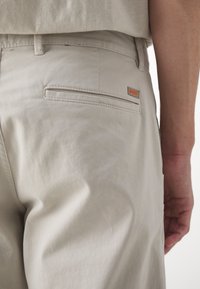 Poche arrière d'un pantalon beige avec une petite étiquette logo orange "BOSS", porté par une personne dont le bras droit repose près du côté.