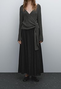 Femme portant un pull enveloppant gris foncé avec un nœud sur le côté, une longue jupe noire et des chaussures noires à enfiler, se tenant sur un sol moucheté.