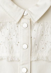 Valkoinen kauluksellinen paita hopeisilla painonapeilla, jossa on kukallinen rei’itetty kirjailu ja hapsureunus rinnuksen kohdalla.