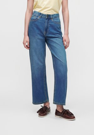 Personne portant un jean bleu taille haute à jambes larges, un haut jaune clair et des mocassins en cuir marron foncé avec des pampilles sur un fond uni.