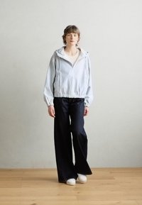 Ribaga kerged jakk kapuutsiga, elastse äärega; koos laia jalgadega tumesiniste denimpükstega ja valgete tossudega puidust põrandal.