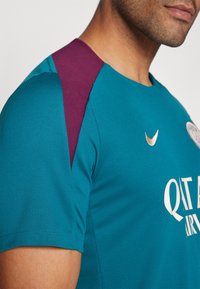Gros plan d'un homme portant un maillot de sport bleu sarcelle avec un panneau d'épaule bordeaux, un logo Nike doré et un texte blanc partiel du sponsor.