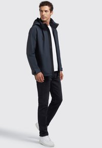 Chaqueta impermeable de color azul marino oscuro con capucha, que cuenta con un cierre de cremallera en el frente y detalles en negro. Combinada con jeans negros y zapatillas blancas.