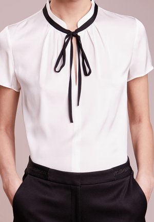Blouse blanche à manches courtes avec détail de nœud noir au col, associée à un pantalon noir taille haute, mains dans les poches, fond neutre.