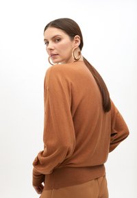 Hnědý svetr s jemnou texturou, volného střihu, s žebrovanými manžetami a detailem knoflíků na ramenech. Doplněno béžovými kalhotami.