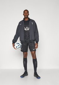 Sort sportsjakke med vertikale striber, sort trøje med "AIA" logo, mønstrede shorts, sorte sokker og fodboldstøvler, mens der holdes en hvid og blå fodbold.