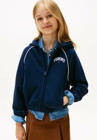 Granatowa bluza z kapturem z białym wykończeniem, z zamkiem błyskawicznym z przodu i logo "TOMMY". Nosi się na jasnoniebieskiej koszuli dżinsowej i w zestawie z brązową spódnicą.