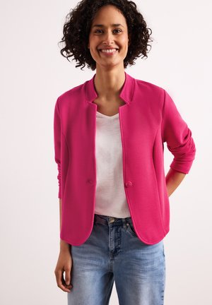 Femme souriante aux cheveux bouclés porte une veste rose vif boutonnée, une chemise blanche et un jean bleu, debout devant un fond uni.