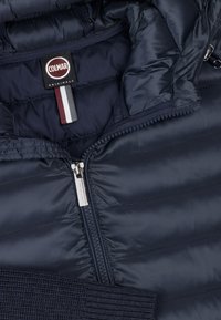 Granatowa pikowana kurtka puchowa o fakturze przypominającej pikowanie, z pełnym zamkiem błyskawicznym i ściągaczami przy mankietach. Posiada metkę z logo i kontrastowy paskowany detal.