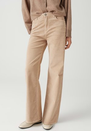 Person trägt beige weit geschnittene Cordhosen, einen braunen Pullover mit Knöpfen und cremefarbene Loafers, steht vor einem schlichten hellen Hintergrund.