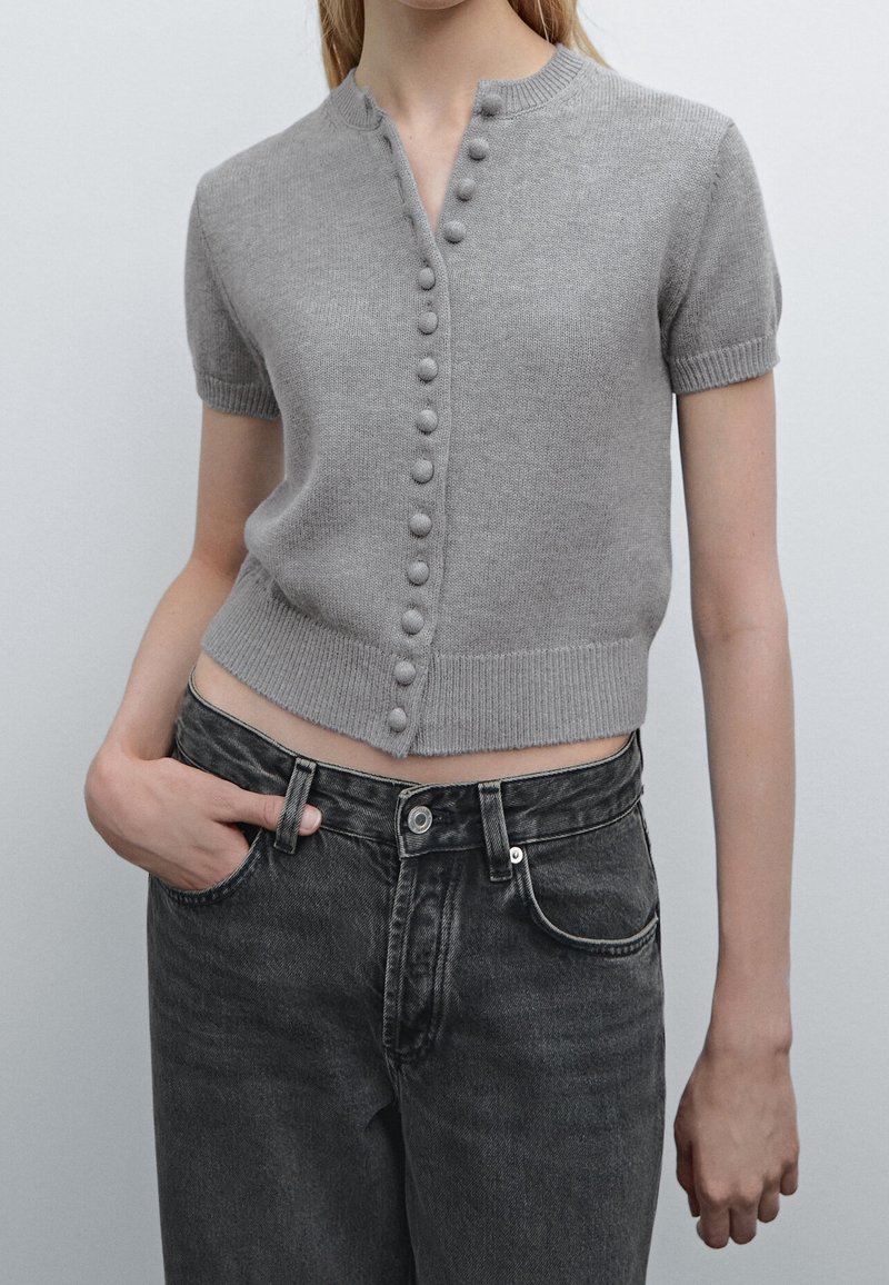 Maglione grigio a maniche corte in maglia con collo alto, dotato di chiusura frontale a bottoni e orlo a coste. Indossato con jeans neri a vita alta.