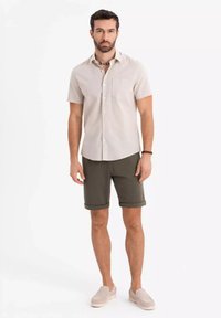 Kortärmad beige skjorta med en ficka, kombinerad med olivgröna shorts och ljusgråa slip-on skor, mot en vit bakgrund.
