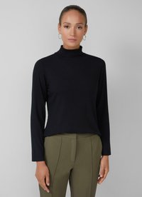 Vrouw in een zwarte geribbelde lange mouw turtleneck en olijfgroene high-waisted maatpakken, staande tegen een lichtgrijze achtergrond.