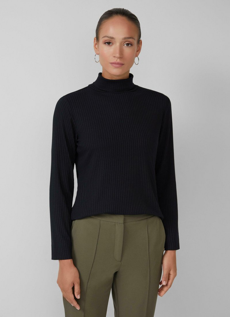 Vrouw in een zwarte geribbelde lange mouw turtleneck en olijfgroene high-waisted maatpakken, staande tegen een lichtgrijze achtergrond.