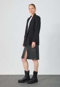 Blazer noir avec une coupe ajustée, associé à une jupe en cuir noir avec une fente sur le devant. Des bottes noires à lacets épaisses complètent la tenue.