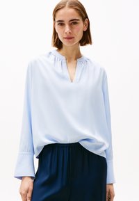 Blusa azzurro chiaro con scollatura a V, colletto arricciato e maniche lunghe con polsini larghi, abbinata a pantaloni blu scuro. Tessuto con una superficie liscia.