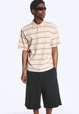 Giovane uomo che indossa una polo beige e bianca a righe, pantaloncini neri e occhiali da sole neri, in piedi con una mano in tasca.