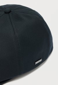 Καπέλο σε ναυτικό μπλε χρώμα με υφή ψαροκόκκαλου, στρογγυλεμένη κορυφή και μικρή μεταλλική ετικέτα λογότυπου στο πλάι. Ραφές με ραμμένα διακοσμητικά στοιχεία κατά μήκος των ραφών.