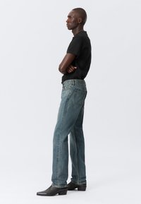 Dark blue denim jeans with a straight-leg cut, featuring back pockets and light distressing, worn with a fitted black T-shirt and black boots.