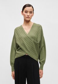 Ζακέτα σε ελιά, με V-λαιμόκοψη, ριγέ υφή και φαρδιά μανίκια. Συνδυασμένη με μαύρες παντελόνες φαρδιές.