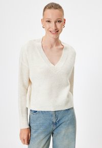 Maglione lavorato a maglia di colore avorio con scollatura a V e trama a costine. Maniche lunghe e design cropped, abbinato a jeans azzurri chiari.